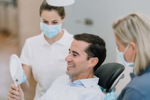 Patient betrachtet nach der Behandlung durch die Zahnärtzin und Zahnarztassistenz seine Zähne im Spiegel